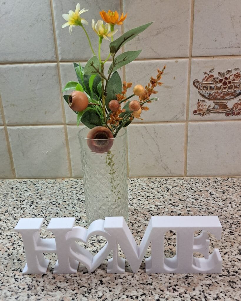 Valkoinen HOME-koriste sydämellä, koristeellinen tekokukkakimppu lasimaljakossa